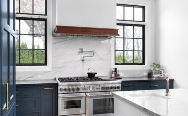 A luxurious kitchen with blue cabinets, a stainless steel stove, granite backsplash and countertop, and black framed windows.